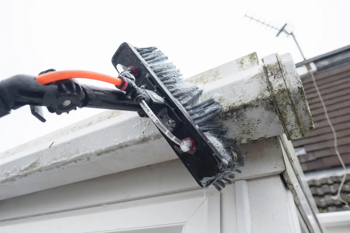 A water-fed brush scrubbing dark buildup from the outside of a white gutter
