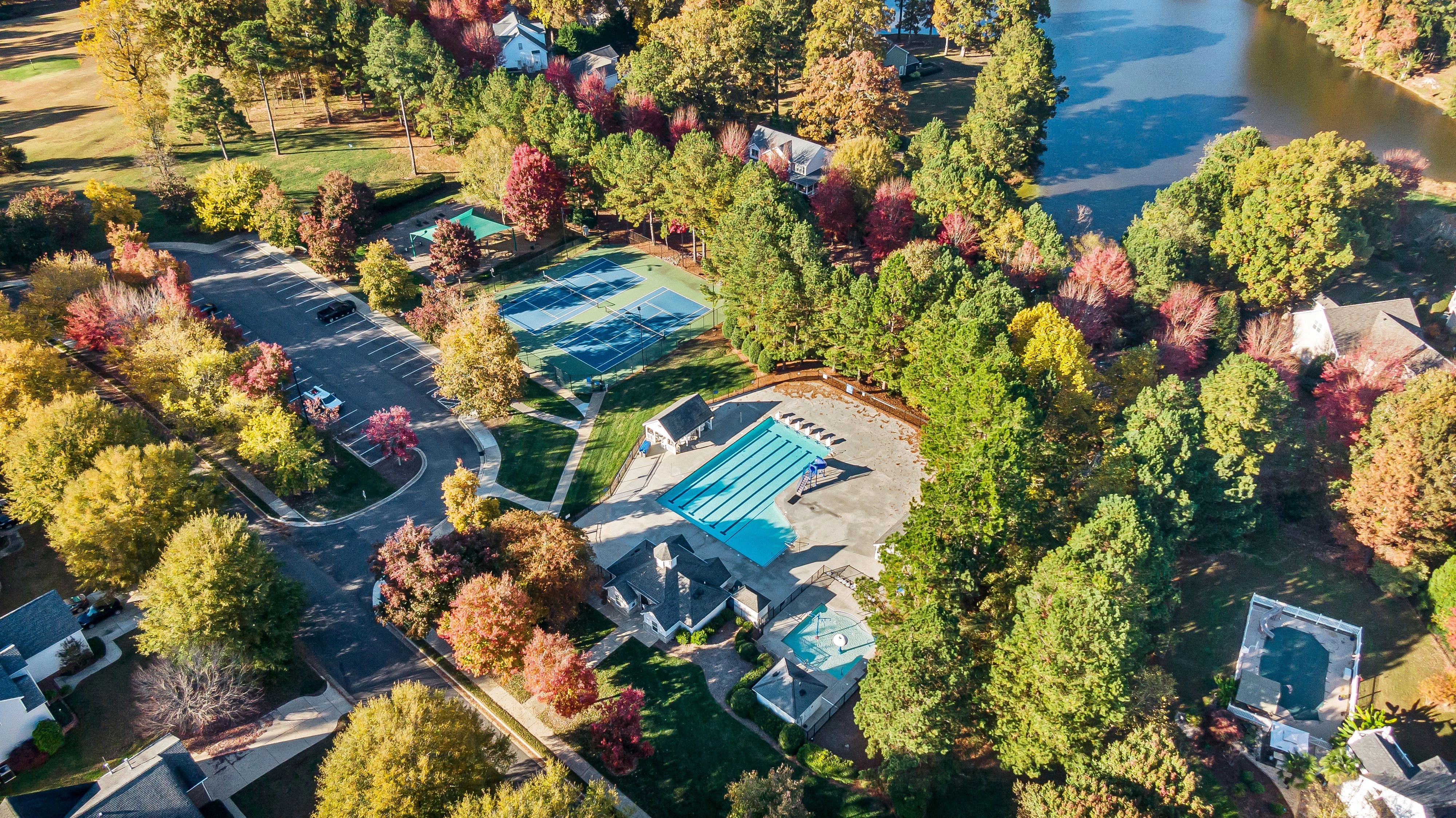 Aerial view of a neighborhood amenity center with a pool, pool house, splash pad and tennis courts among autumn trees beside a lake