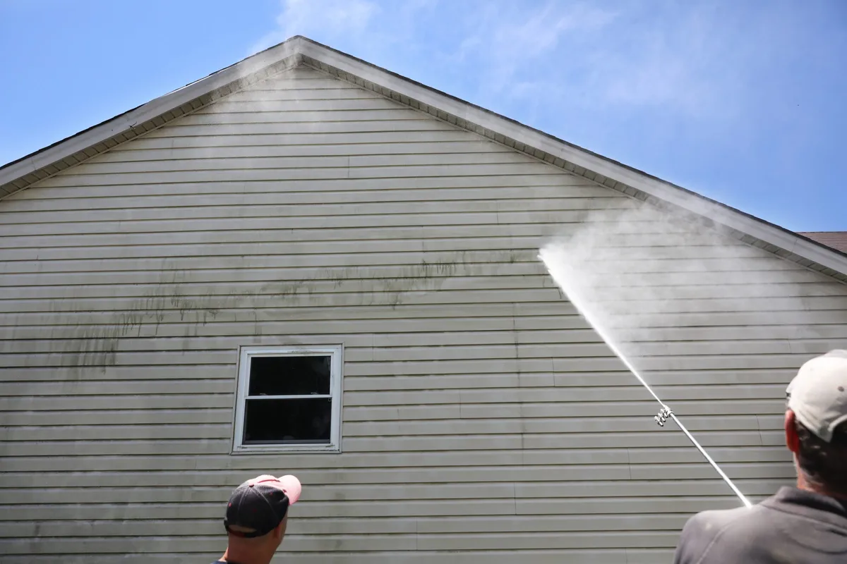 A worker rinsing streaked horizontal siding beneath a house gable