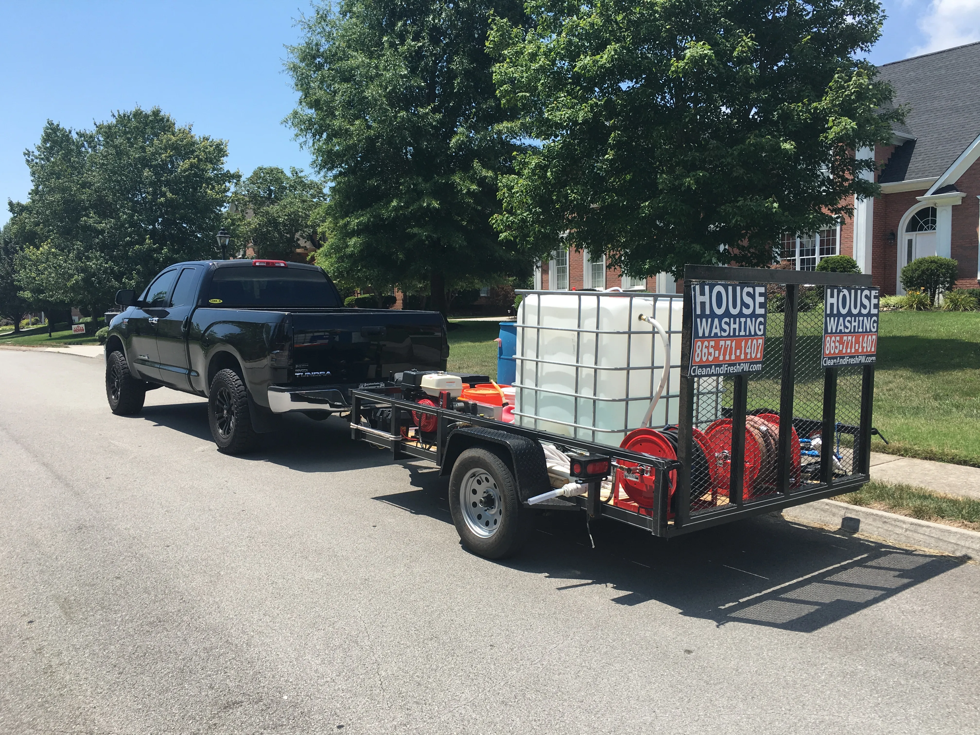 A black pickup towing an open utility trailer loaded with a water tank, a pressure-washer engine, red hose reels and House Washing signs