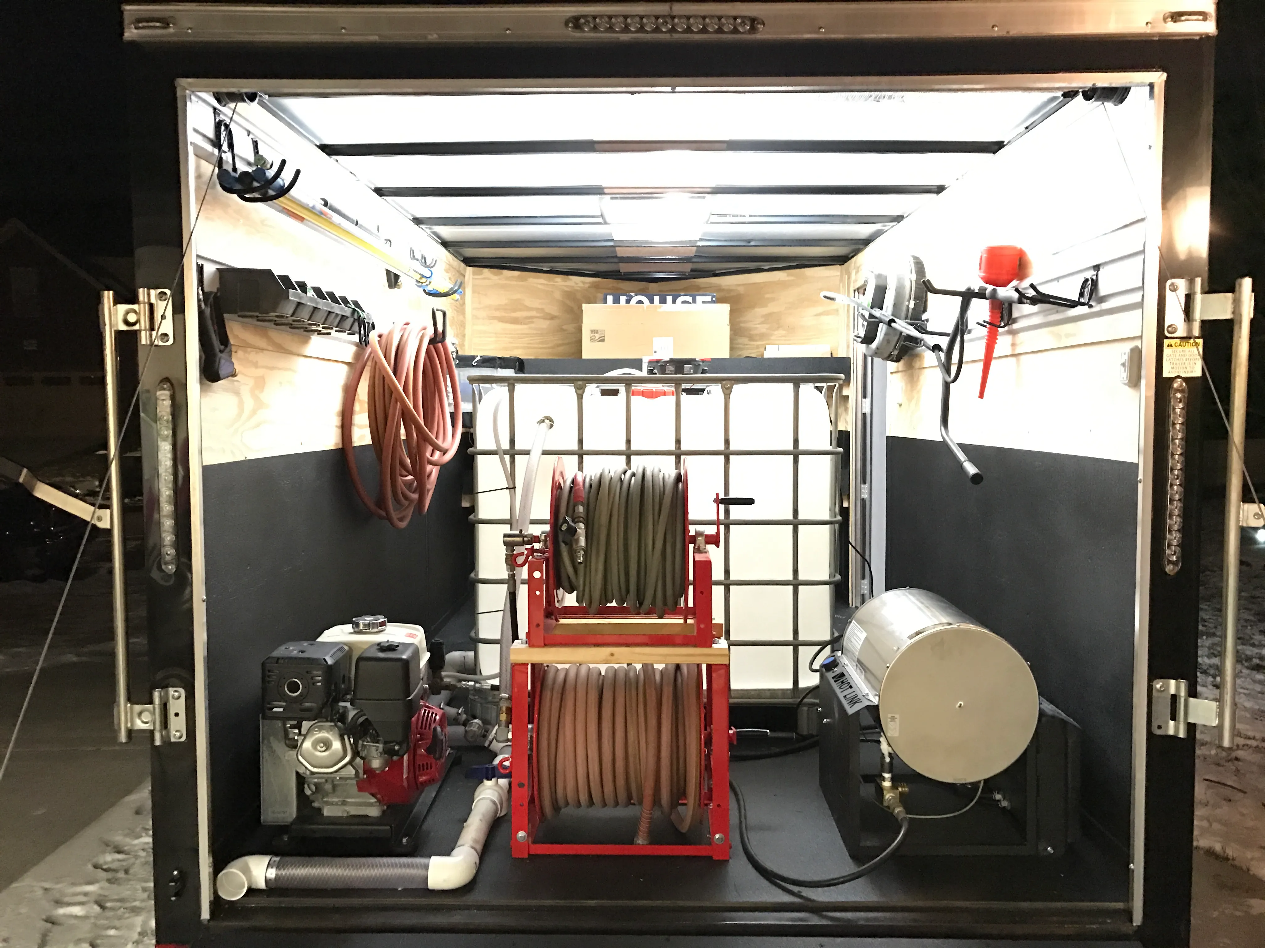 Inside the enclosed trailer at night: a pressure-washer engine, stacked hose reels, a water tank, a hot-water unit and hoses and wands hung on the walls