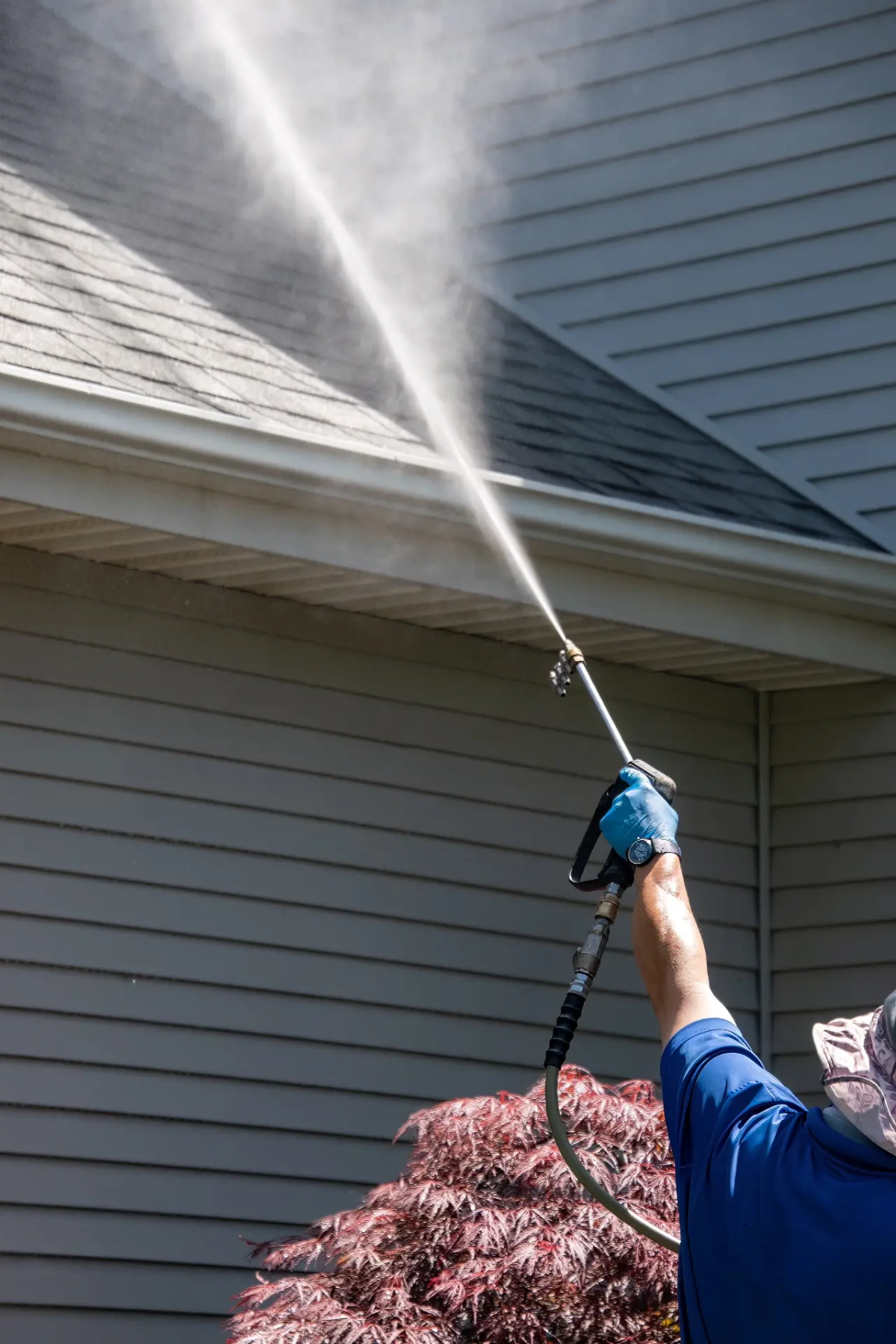 A worker holding a spray wand toward the siding and eaves of a house