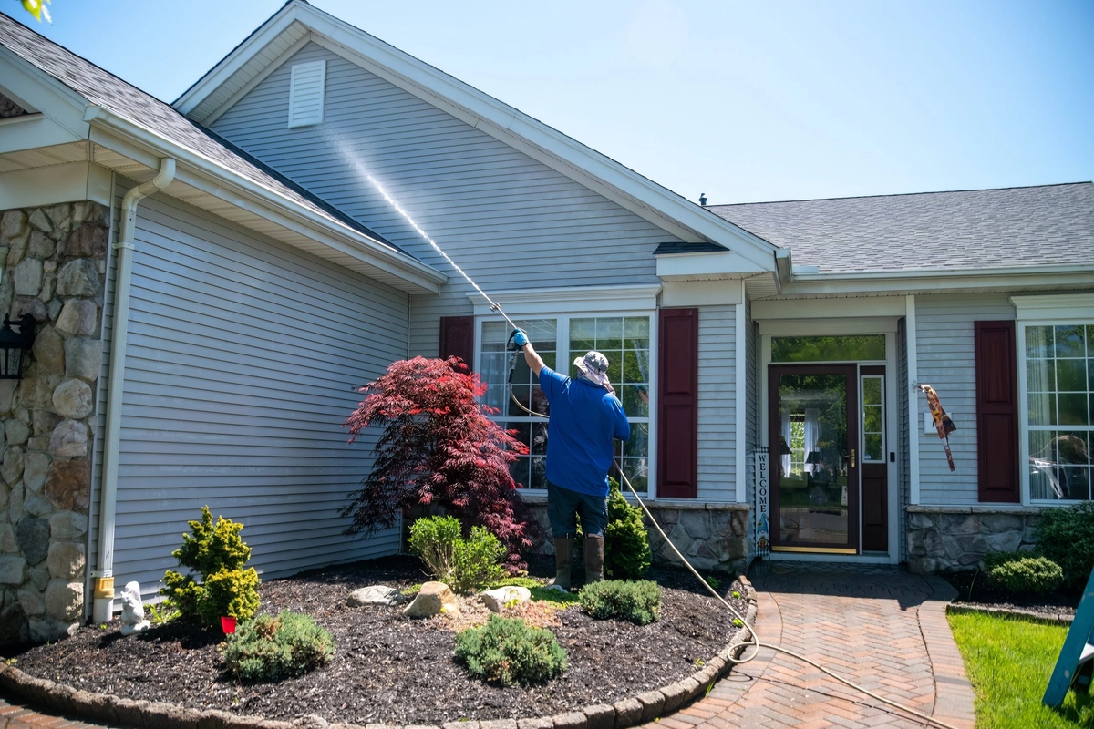 A worker spraying the upper siding of a house from the ground beside planting beds